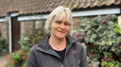 Christine Hellio is a woman with short blonde hair. She is wearing a grey fleece with a black top underneath and a chain necklace. The background is an out of focus granite cottage with plants all over it.