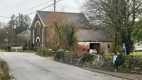 Road near Caer Salem Baptist Chapel. The chapel is visible and trees are in the background. Police tape is also visible across a driveway.