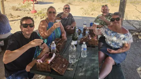 A group of five people sat at a picnic table in the Gran Canyon. They are all looking at the camera smiling and holding a drink aloft.