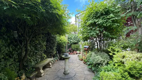 Ann Lush An image of one area of Ann's garden. It has lots of greenery, and a centre piece of a bird bath, stone tiling on the floor with a small bench.