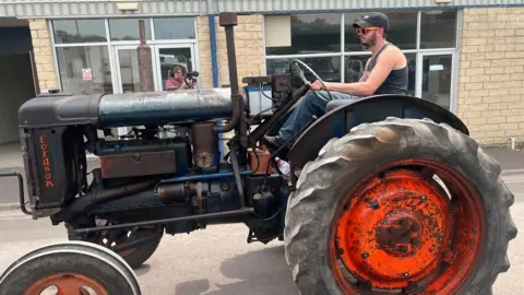 A man is riding a vintage tractor and wearing a cap and sunglasses.