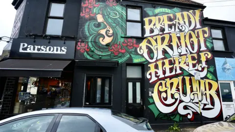 UPFEST A large artwork filling a two-storey building reads "bready or not here I crumb" in stylised, curly capital letters. A carton image of a woman with flowing green hair is next to the writing. Another part of the building is a bakery shopfront with a sign saying "Parsons". 