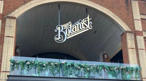 A gold sign for The Botanist hanging from a brick archway over a glass balcony with fake greenery