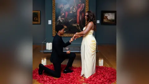 Emmanuel Dosumu/adidolvisuals A man in an all black suit is down on one knee in front of a large painting. A woman in a cream dress stands smiling at him as he takes her hand. They stand among a pile of rose petals with several candles lit on the floor next to them.