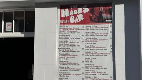 A poster outside a building lists upcoming live music events at The Marr’s Bar, with dates and tribute acts shown.
