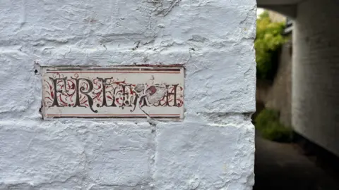David Parr House A hand-painted rectangular sign on the corner of a white-painted brick wall. It is a little battered but it is possible to see it says FR Leach in brown cursive letters with red patterns painted around the letters. 