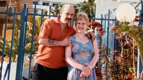 Hutchinson family Tony Hutchinson and Wendy Hutchinson pictured on holiday. Wendy is wearing a blue dress with floral patterns, with Tony in an orange top and dark trousers. They are stood by blue iron gates and are surrounded by exotic plants. 