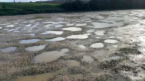 Chris McHugh/BBC An outdoor gravel car park is seen almost completely covered in circular, rain-filled potholes. A grassy bank with a road behind it can be seen in the background