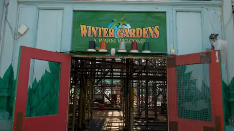 Qays Najm/BBC The front of the Winter Gardens in Great Yarmouth. A sign sits above the doorway in yellow lettering which says, WINTER GARDENS A WARM WELCOME. The red doors are wide open exposing a structure inside being supported by poles.