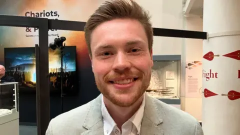 BBC/Seb Cheer A man, Harry, wearing a light-coloured suit, looks at the camera and smiles. He is standing in the exhibition.