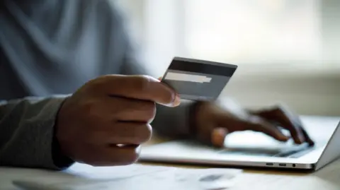 A man wearing a grey sweater sitting at a desk in front of a laptop holds a credit card in one hand between his index finger and his thumb, while his other hand is tapping the keys on the laptop. We cannot see the face of the man.