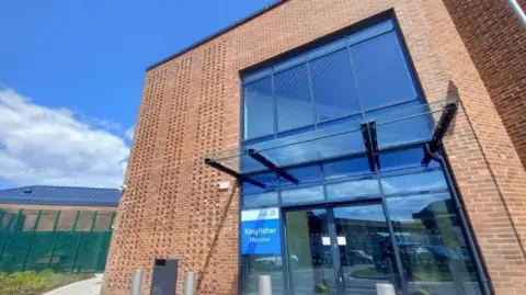 Derbyshire Healthcare NHS Foundation Trust A new brick building with a large glass entrance, with green fencing in the background
