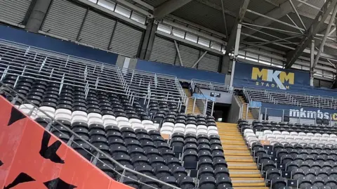 An empty sport stadium stand with rows of black and white seats. Yellow steps form a walkway between blocks. Advertising hoardings are visible. To the left, a wall has been painted orange and black to resemble tiger stripes.