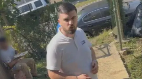 Ginia Hines A man with short dark hair and a neatly trimmed beard is wearing a white t-shirt. He is standing on a small pathway in the front garden of a home, with another man in work clothing sitting behind him eating. The second man's face is blurred. Cars, a hedge and a small patch of grass are visible in the background. 