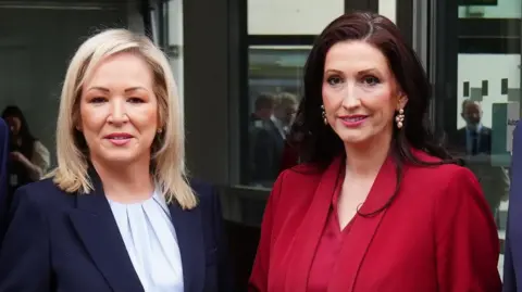 Michelle O'Neill, a woman with shoulder-length blonde hair, and Emma Little-Pengelly, a woman with long, dark hair, pose for a photo. O'Neill is wearing a navy suit jacket with a pale pleated-neck top and Little-Pengelly is wearing a bright red suit jacket over a matching red blouse. 