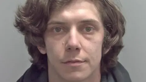 A police custody image of a man staring blankly at the camera. He has thick wavy brown hair and hazel eyes. 