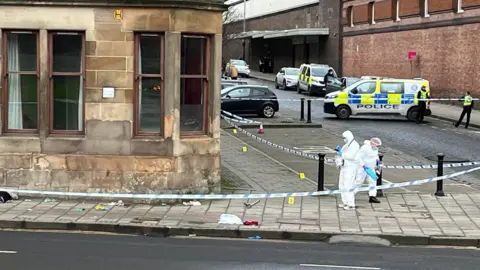 Two forensic officers, wearing white hazmat suits look for clues at the  scene of a disturbance in the west end of Glasgow. They are working on the pavement behind an area cordoned off with blue and white police tape. Two police officers are visible in the background next to a Police Scotland van. A second police van is parked nearby 