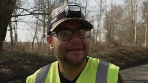 Shaun Whitmore/BBC A man in glasses and hi-vis vest with a baseball cap and camera on his head