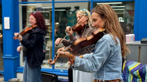 Grainne is on the right playing the violin outside a shop, with two others playing as well to the left. She has long brown hair and a denim jacket. The is a shop front behind them with bright blue walls and large windows.