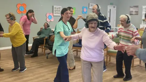 BBC A group of older people and nurses dancing in a room. The older people are wearing headphones. 