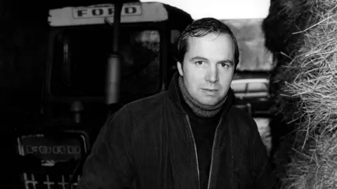 Charles Collingwood, wearing a corduroy coat, dark jumper and scarf, stood in front of a tractor, with hay bales on the right of the picture 