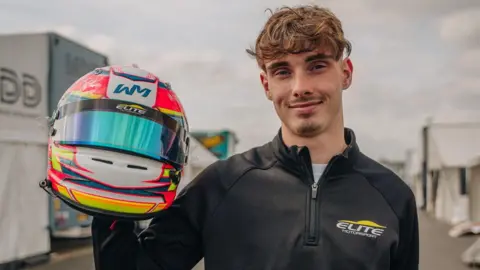 Instagram/wilmac.7 A young man with a light beard is holding a racing helmet and smiling towards the camera
