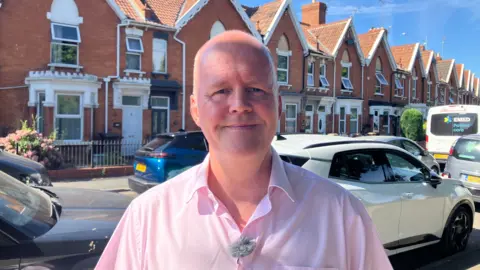 Ashley Fox wearing a pink shirt and smiling at the camera with houses on Cranleigh Gardens behind him