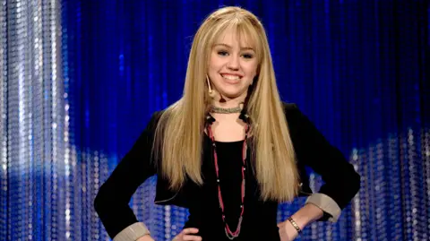 Getty Images Miley Cyrus as Hannah Montana. Hannah is a teenage pop star, she wears a long blonde wig and black jacket over a black top, posing with her hands on her hips. 