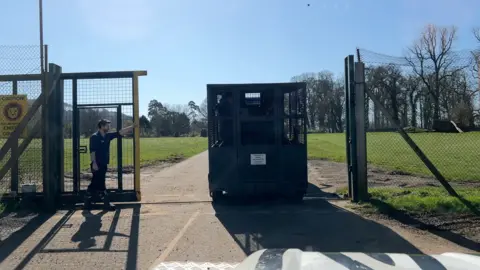 The meat wagon is driven through the security fence, pulled by a tractor. A keeper holds the gate open.