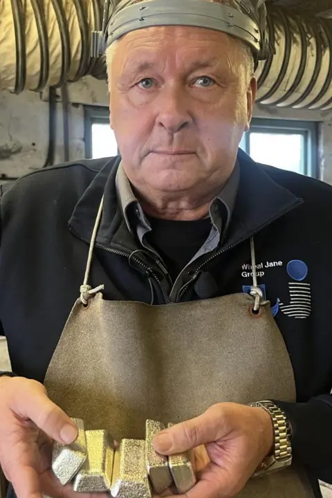 An older man with grey hair looking straight at the camera, wearing a face visor pushed back on his head, a black sweatshirt and a brown apron. he is standing in a shed with a big pipe behind him and is holding up six shiny tin ingots to the camera