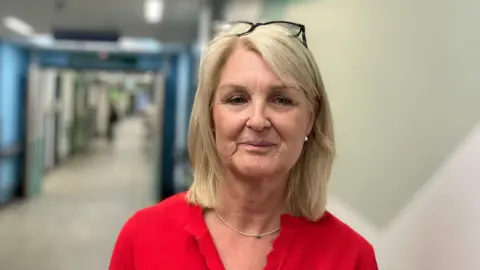 Vikki Irwin/BBC Kirsty Rowden is standing next to a wall on the right. She has shoulder length blonde hair and has black framed glasses resting on top of her head. She is wearing a red shirt, silver necklace and silver earrings. 