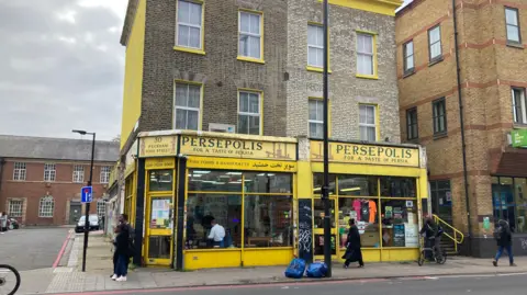 Exterior view of Persepolis, a Middle Eastern shop and restaurant. The shop has a yellow exterior. People walk past on the street. 