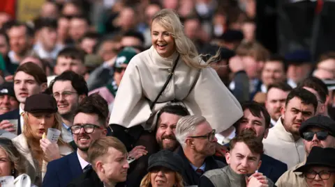 PA A crowd of people in jackets and hats looking in one direction. One man is grimacing as he carries a woman in a white cape-style jacket on his shoulders.