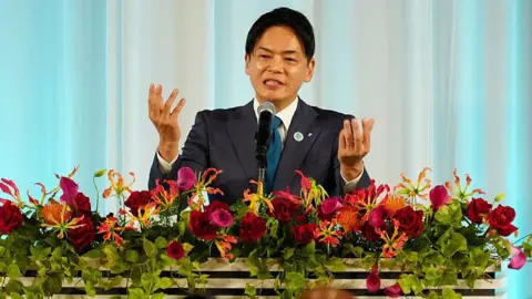 Takeharu Yamanaka in a suit addresses an audience from a decorated podium filled with vibrant flowers against a curtain backdrop