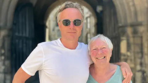 Shariqua Ahmed/BBC Toby, a man with small grey hair, wearing a white T-shirt. He is standing next to Pauline, a woman in a green T-shirt, smiling for the camera.