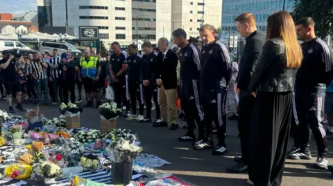 Newcastle United head coach Eddie Howe stands with his head bowed outside St James's Park with club staff, players and fans. There are floral tributes on the ground at their feet.