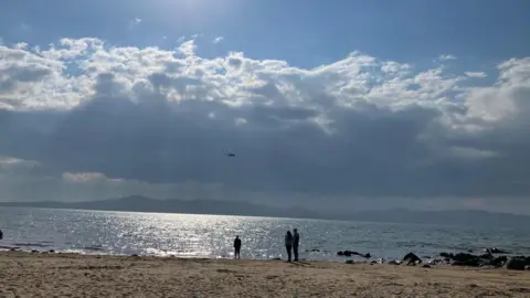 Three people standing on Buncrana beach in County Donegal are looking out to sea on a sunny day and in the sky a coast guard helicopter can be seen searching the water for swimmers who have been reported missing