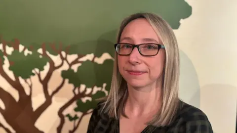 A woman stands in front of a picture board. She is wearing a dark shirt and has glasses. Her hair is long and straight. She is smiling. The board behind her shows part of a picture of the trunk and branches of a hawthorn tree. 