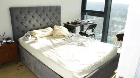Met Police The bedroom of the house where Zhenhao Zou lived. There is a grey bed with a white sheet over the mattress. To the right of the bed is a desk and chair and to the right the window overlooks London