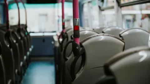 A wide shot taken of an empty bus. It shows a number of grey plastic seats with grey leather covering the seats. There is a blue walkway and a number of pink poles with stop buttons on them.