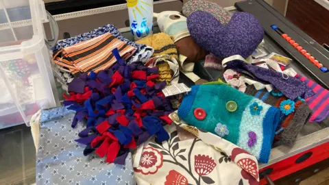 A table filled with colourful, soft, and patterned fabric creations. The items appear to be part of a crafting session or display. Several stacks of rectangular cloths with patterns such as stripes, stars, and flowers. There are several multicoloured, textured knit items in various shapes. There is also a stuffed fabric heart and a pale lantern.