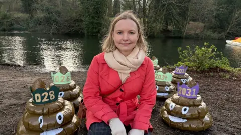 Martin Heath/BBC Kat Hobbs with long blonde hair, wearing a red coat and dark trousers. She is sitting with poo swirl emojis on either side, which have coloured hats showing different years.