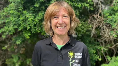 A smiling woman with short blonde wavy hair in a black button-necked T-shirt with a green T-shirt underneath