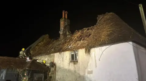 Wilton Fire Station Two firefighters wearing uniforms and yellow helmets climbing on the roof of a building adjacent to the burnt property. The thatched roof is almost completely gone, except a portion on the right which could be salvaged. It is night time and the sky is completely black, with spotlights shone on the house.