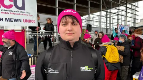 Deio Owen pictured in front of the Senedd in a black zip-up NUS Cymru jacket and a pink UCU hat, wearing a backpack.