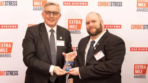 Combat Stress On the left is Admiral Sir Philip Jones who has silver white hair, and is wearing a white shirt, black suit jacket and black tie. He is holding on to the one side of a glass trophy. On the right is Pete White, who has very short brown hair and facial hair. He is wearing a white shirt, blue tie and black suit jacket. He is holding the other side of the glass trophy