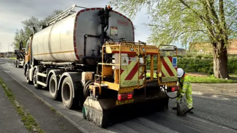 A highway maintenance vehicle on a road with a man wearing a white hard hat and a fluorescent outfit holding a pipe into a bucket