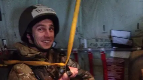 Collect/PA Real Life Rob dressed in his RAF uniform on board a plane. He is smiling at the camera and wearing a black helmet, sitting and holding a black handle attached to a yellow strap. 