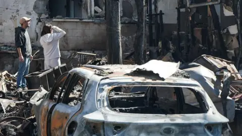 Reuters A couple look at badly damaged building, with a burnt-out car in the foreground, in Nahariya, Israel (17/03/26)