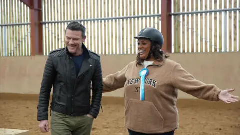Luke Evans and Alison Hammond are inside a horse stable. She is wearing a helmet and a blue ribbon.